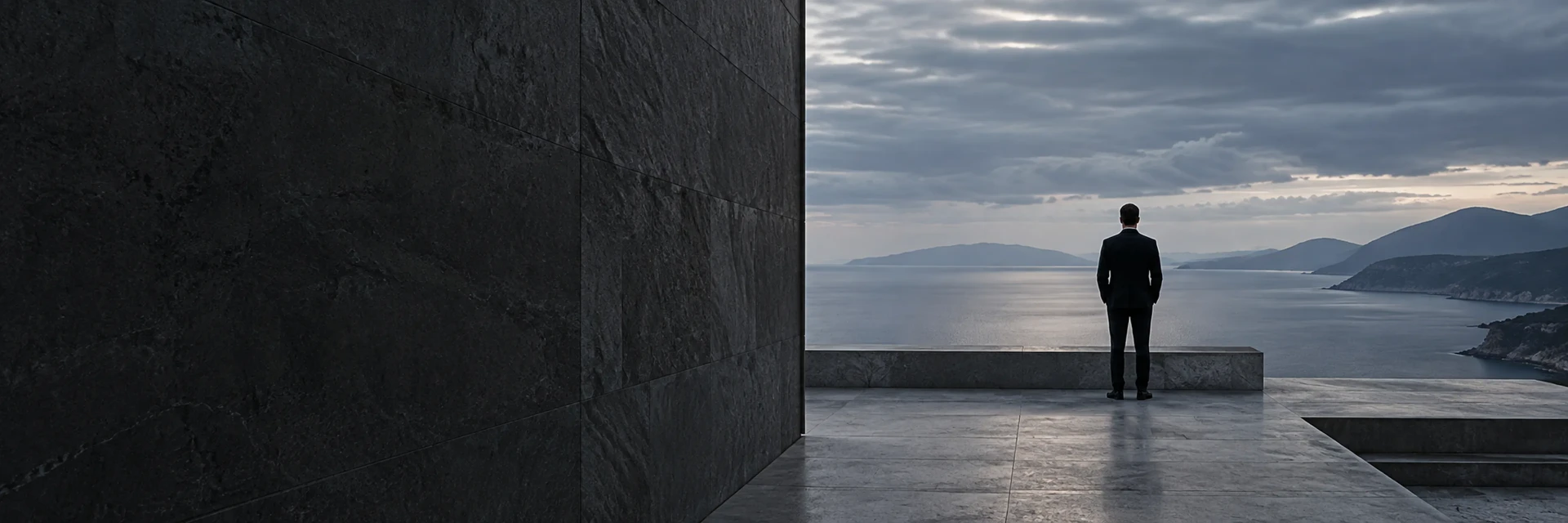 A figure on a stone terrace looking out over the sea and a distant coastline.